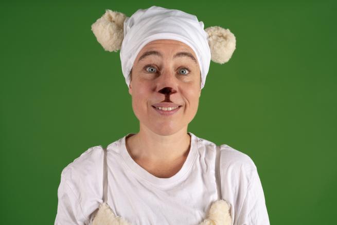 A caucasian woman with blue eyes dressed as a white sheep looks directly at the camera. There is a green background