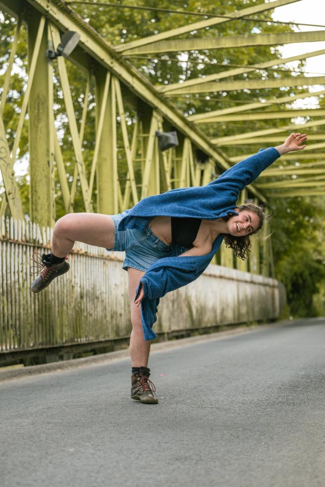 Dancer in shorts and a blue shirt, standing on one leg on a bridge with yellow metal across it. Industrial background, with the dancer in the middle of the frame with her right leg outstretched and left arm stretched above her head.