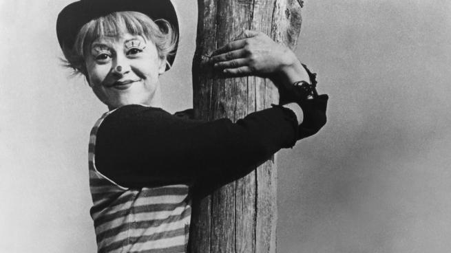 The protagonist, Gelsomina, adorned in clownish makeup, hugs a large wooden pole while smiling at the camera.