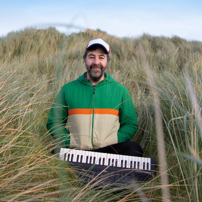 David O'Doherty stood in a long grass wearing a green and beige jacket with a keyboard.