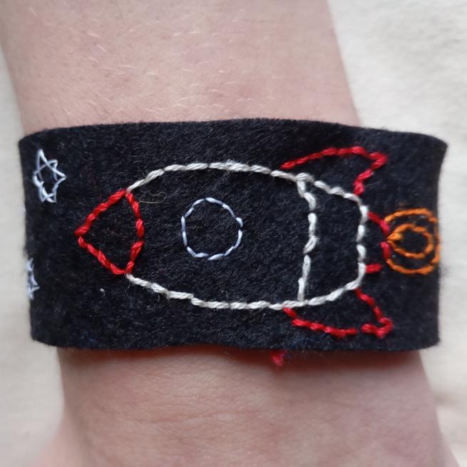 a wrist showing black fabric wristband with a stitched rocketship