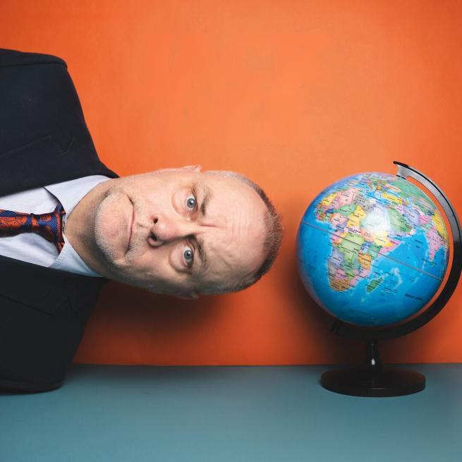 Shoulders and head of Jack Dee wearing a suit lying on his side next to a globe. 