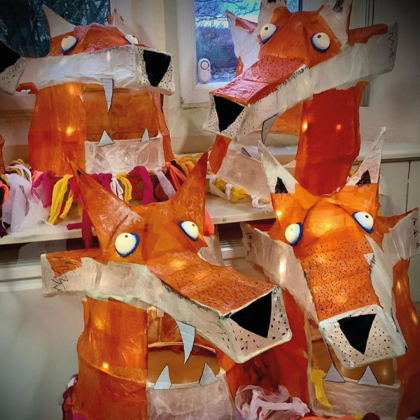 The image shows brightly coloured headdresses in the shape of fox heads. The headdresses are lit with fairy lights and are made out of willow and paper so the light shines through.