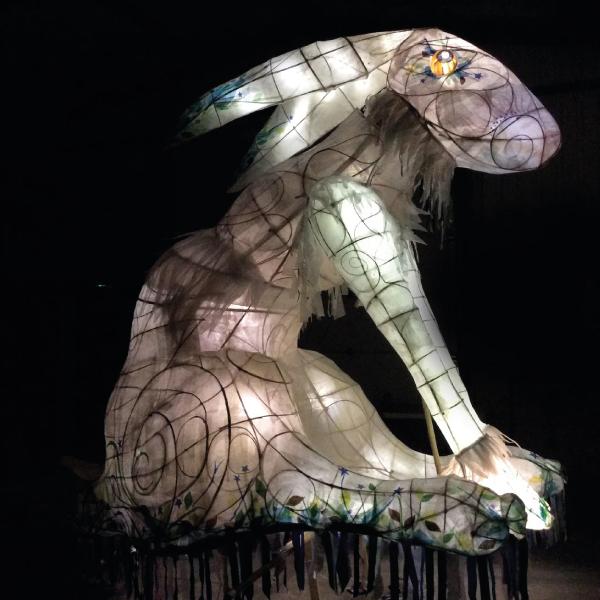 The image shows a large-scale lantern in the shape of an enormous hare, the lantern is made out of willow and paper. The lantern is lit from the inside with fairy lights and shines bright against a dark background.