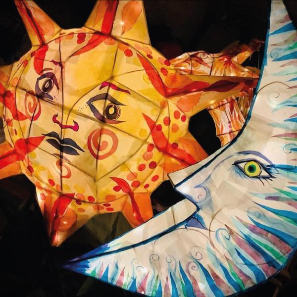 The image shows to brightly coloured lanterns made out of willow and paper. The lanterns are in the shape of a sun and moon, both have faces and are lit from within using fairy lights.