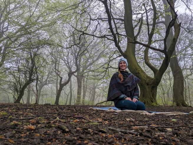 A photograph of Alice sat on a yoga mat within woodland. 