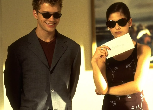 Young man and woman both wear sunglasses and stand next to each other. The man wears a blazer and grins. The woman wears an evening dress, pouts and is holding a white envelope.