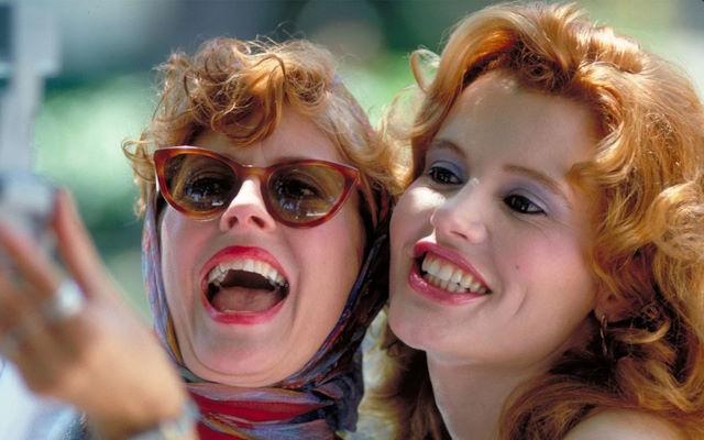 Two women wearing lip gloss smiling, one wearing sunglasses.