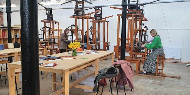 Image Shows the weaving shed- filled with workshop participants working on traditional dobby looms- in the foreground is a large table with a vase of daffodils. 