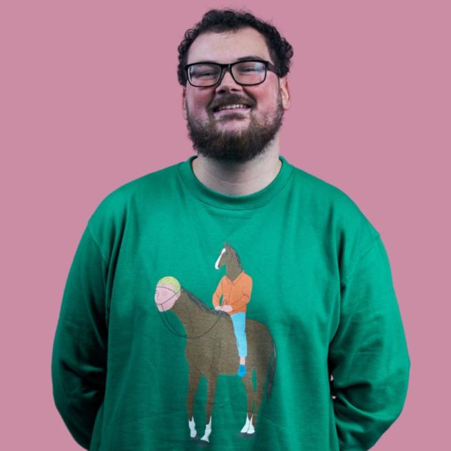 Alex Mitchell smiling, wearing glasses in a green Bojack Horseman sweatshirt where a man with a horse's head is riding a horse with a human head. Alex is stood against a pink background.