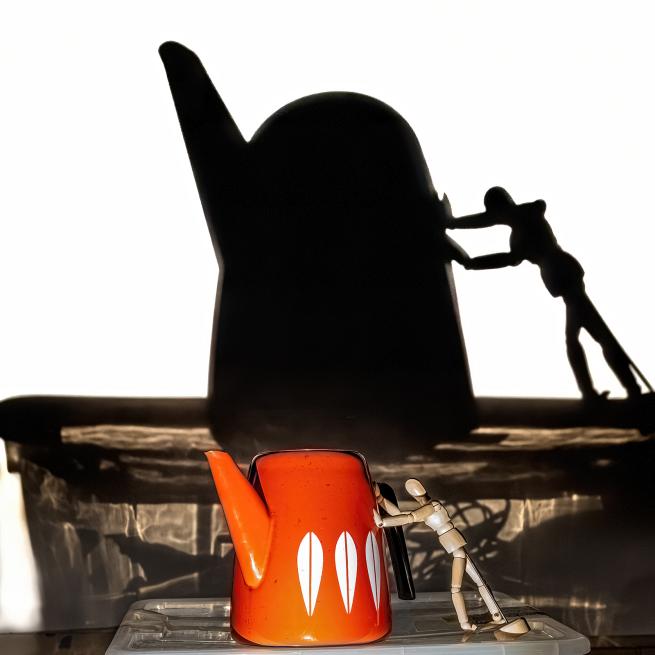 Photograph of a small wooden mannequin pushing an orange 1970s coffee pot. The photograph is lit so that a larger shadow of the mannequin and coffee pot appears in the background.