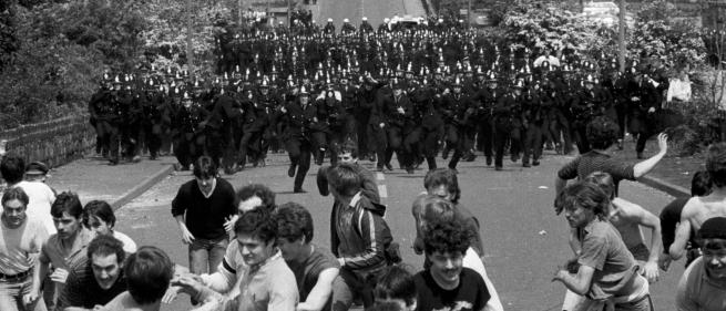 A group of men in the foreground run away from an even larger group of police in the background.