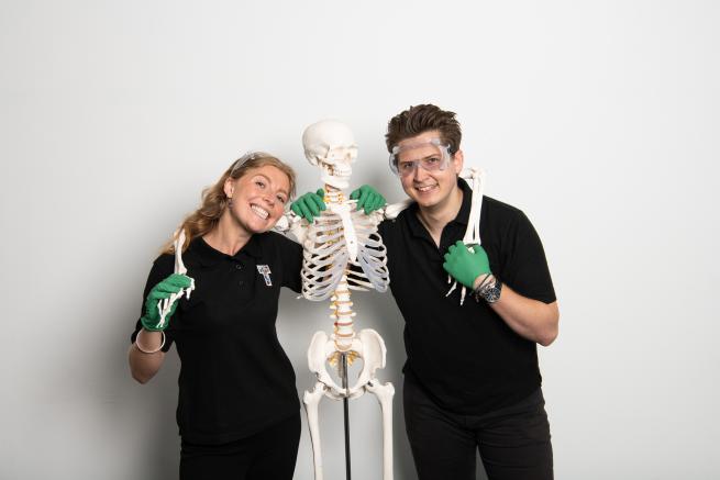 An image of two people wearing surgical gloves with their arms around a model skeleton.