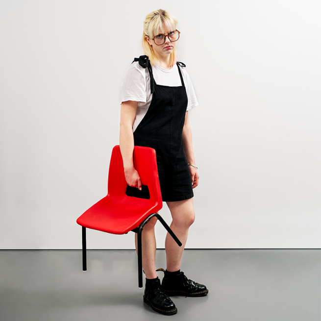 Women with a whit et-shirt, round glasses, a black jump suit and black shoes holding a red small children's chair as she looks to the front. 