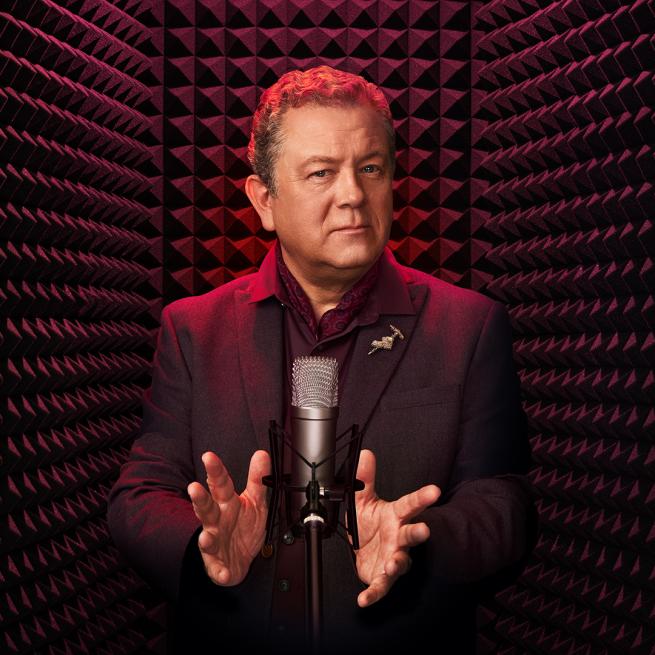 Jon Culshaw in a dark maroon suit stood at a recording microphone in a recording booth.