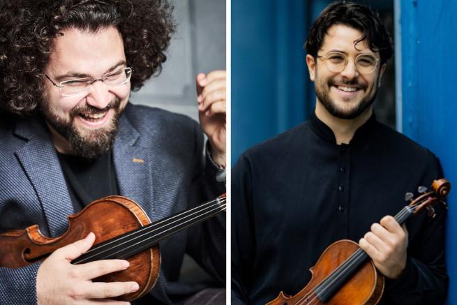 There are two white men holding violins in the foreground. One has a lot of dark curly hair, glasses and a beard, and is wearing a dark grey blazer. The other has shorter dark hair with a black shirt. They are both smiling. 