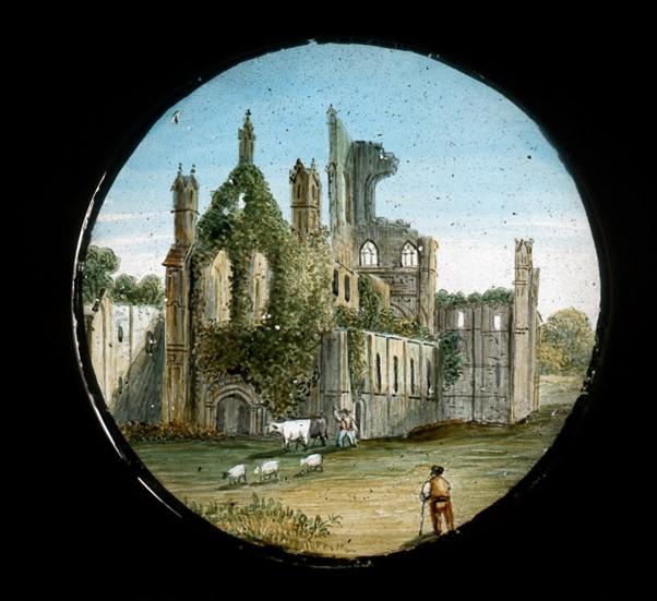 A lantern slide featuring a painting of monastic ruins of Kirkstall Abbey. In the foreground are some sheep and a man, presumably a shepherd, looking back towards the ivy covered Nave of the Abbey.