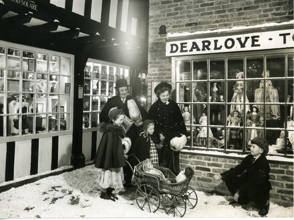 A black and white snowy street scene in a Victorian museum. A family dressed in period costume are set against the backdrop of Abbey House Museum's street as it appeared in the 1950s and 60s, with fake snow on the ground.