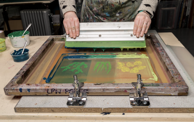 Image shows a screen with a design and green ink on it, with some hands pulling a squeegee across the screen