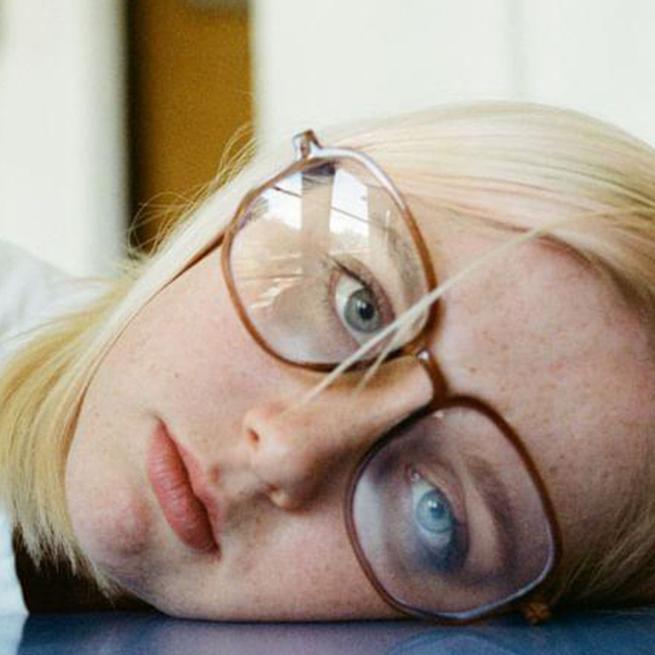 Close up on the face of a young girl wearing glasses, her head is laying on a table as she stares into the camera with a fed up expression. 