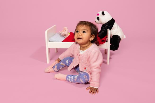 An image of a child in front of a pink background with a small bed and a cuddly panda toy.