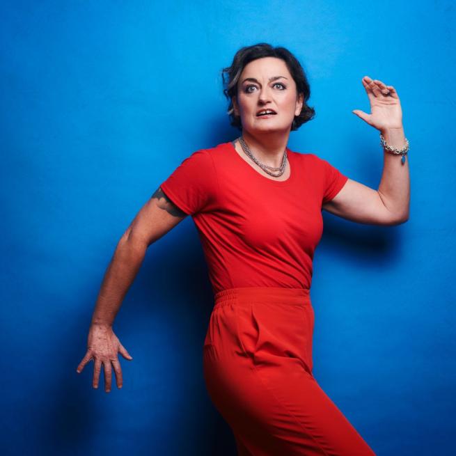 Zoe Lyons in an all red outfit against a blue background with her arms shaped as if she's running with her left out front bent up at the elbow and her right behind her bend down at the elbow.