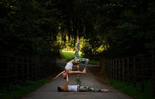 2 women in a park pathway, covered by trees. Theyr'e wearing yoga clothing and are balanced, one holding up the other in the air, who is arched back and left leg up in a striking pose