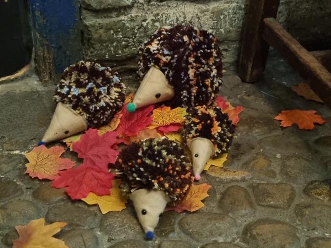 A small family of fabric hedgehogs sits on a pile of leaves in a mock up Victorian backstreet