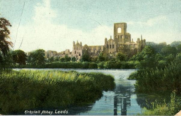 A picture postcard featuring Kirkstall Abbey - the monastic ruins are reflected in the water and there is grass surrounding the river which is in the foreground