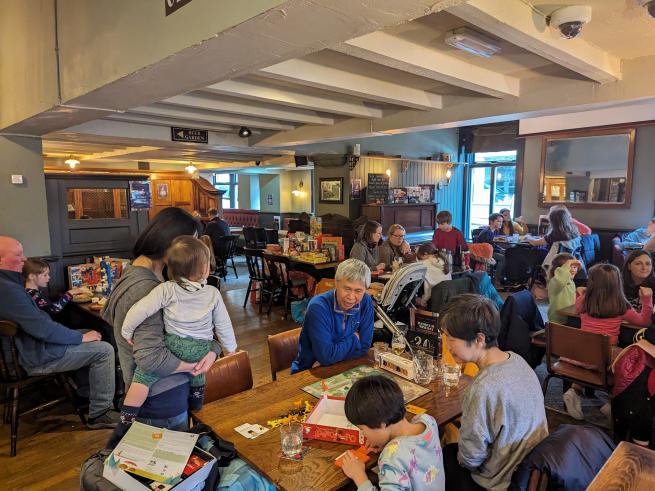 People sitting at tables playing games. In the foreground one person is standing holding a baby whilst playing. 