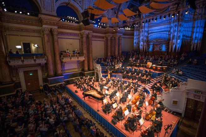 A grand Victorian concert hall with lots of members in the audience in front and behind of the stage. There is an orchestra on stage and a grand piano.