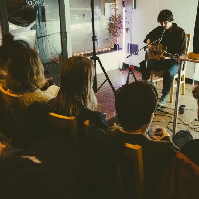 Musician Chris Brain sits in a small and intimate performance space, playing an acoustic guitar. The photo is taken from just behind a small audience that sits just in front of Chris, listening intently. 