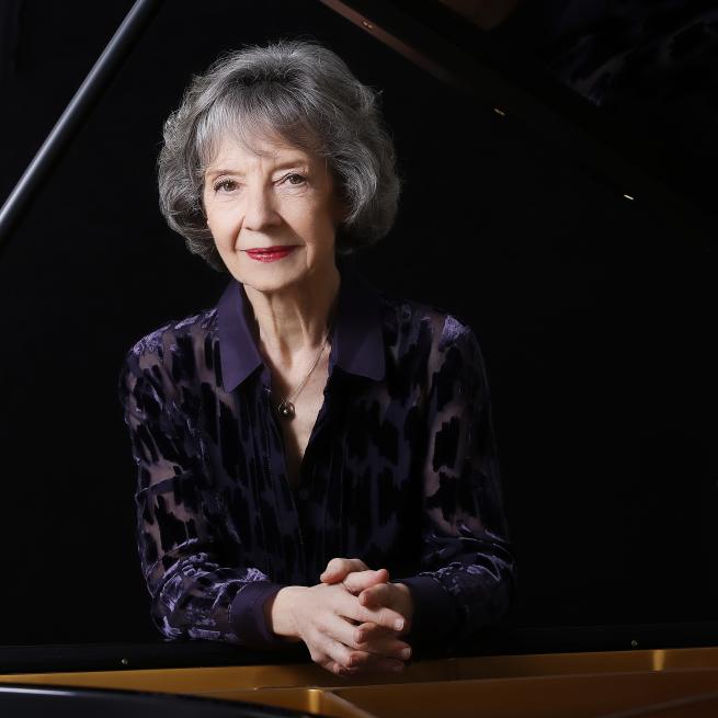 A white woman in her late 70s leaning on a black grand piano with her hands crossed. She is wearing a dark purple and black shirt with red lipstick. She has grey short hair.