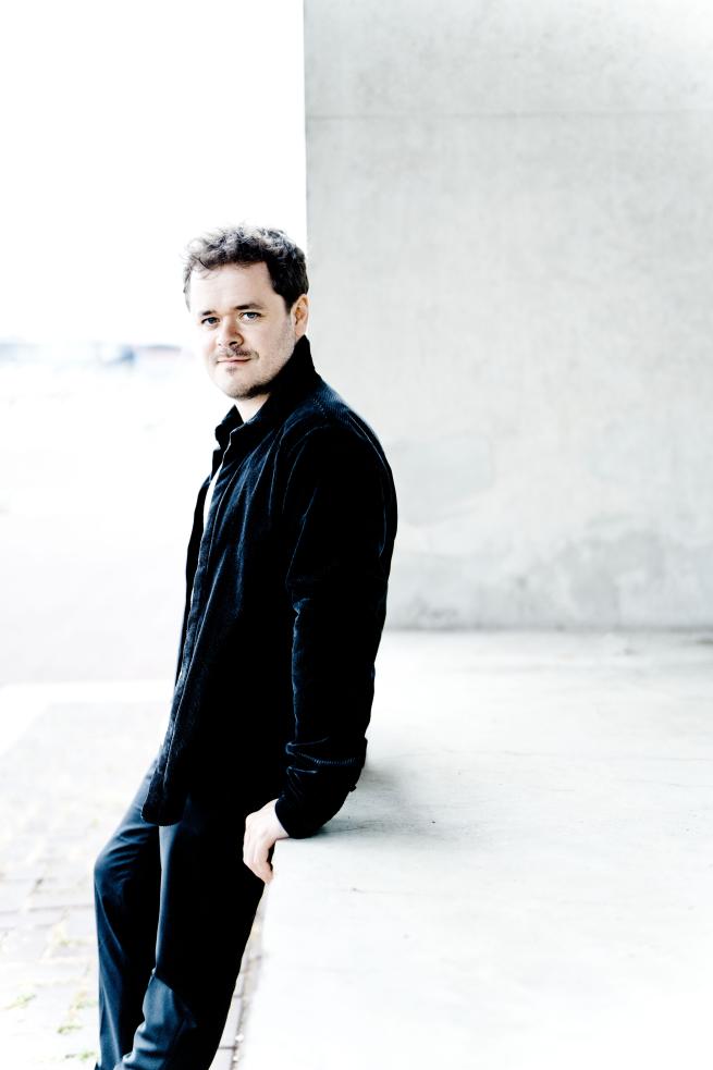 A white man in his early 30s wearing a dark coloured shirt, back trousers and a white t shirt. He is leaning against a white hip-height wall and is smiling at the camera.