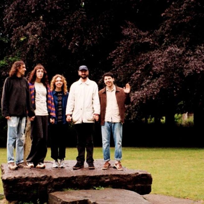 The five band members of Nature Kids stand on a large rock in a park. One member of the band is raising his left hand to wave at camera. 