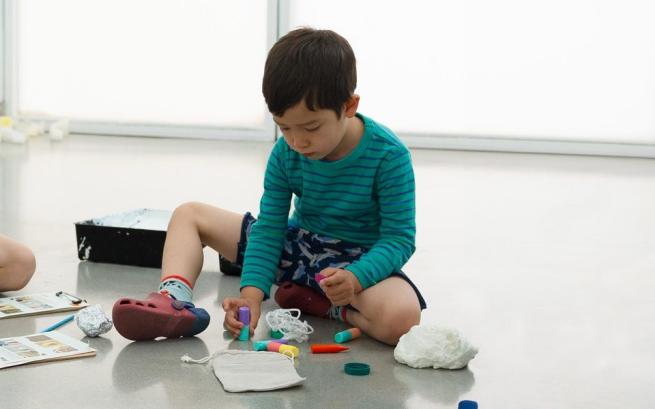 A child sits on the gallery floor surrounded by activities including building blocks, string and crayons.