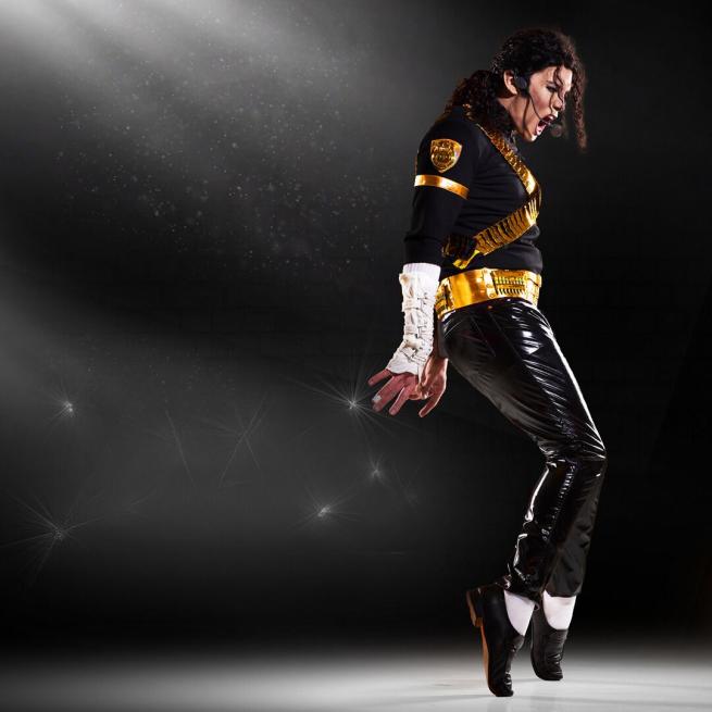 A Michael Jackson tribute act wearing a black and gold-detailed top, white wrist warmers and black latex trousers. He has a headset microphone and is up on his tip-toes with a light shining on him