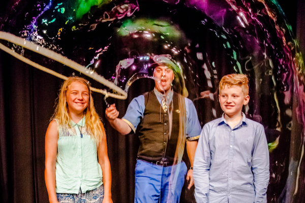 Two children stand either side of  a man with a large bubble wand, he is producing a large tube shaped bubble to cover them both. 