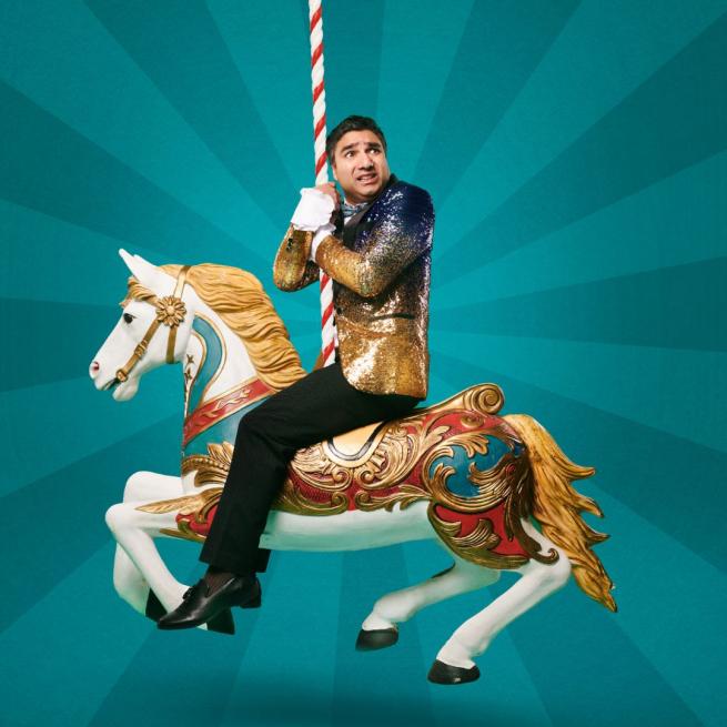 Nick Mohammad wearing a sequinned navy and gold suit jacket with black trousers riding a merry-go-round horse looking scared. He is against a dark teal / light teal patterned background.