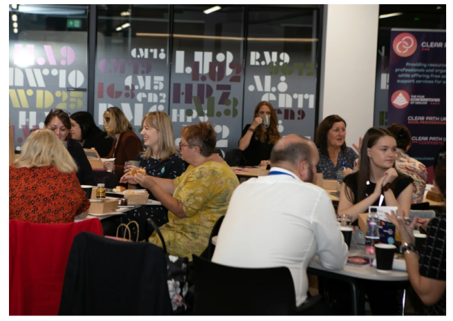 Our 2022 attendees sitting at tables in a modern room, eating and talking. Some are holding drinks and snacks in hand, while behind them is a glass wall with a graphic design and an informational poster.