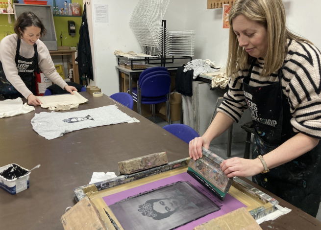 Two women screen printing a portrait of a woman on to blank canvas tote bags.