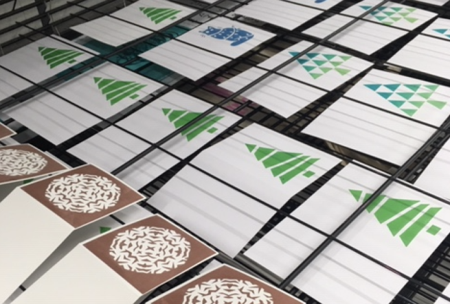 Screen printed Christmas Cards on a drying rack, featuring images of snowflakes, Christmas trees and cats, in blues greens and browns.