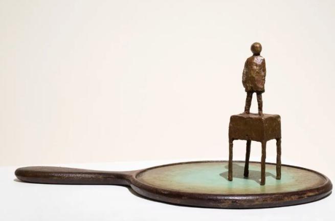 bronze statue of a child stood on a small desk, that looks like its stood on a old fashioned hand mirror