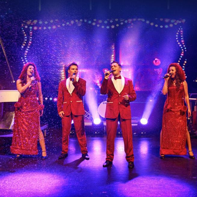 A group of four singers in sparkly red outfits singing into handheld microphones with a purple background.