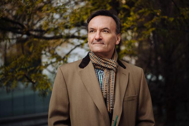 A white man in his early 60s wearing a camel coloured coat with a black collar and a orange, black and white printed scarf. He is looking towards the left and there are trees in the background. 