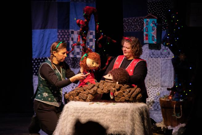 Two people controlling a puppet of a little girl, and a hedgehog stand on a stage with a patchwork backdrop