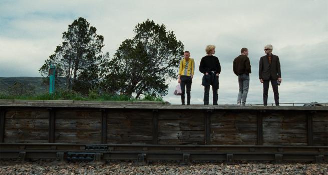 A still from Trainspotting featuring four characters (played by Ewen Bremner, Kevin McKidd, Ewan McGregor and Jonny Lee Miller) stood on a train station platform.