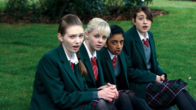 Four girls in school uniform sit next to each other outside and look shocked.