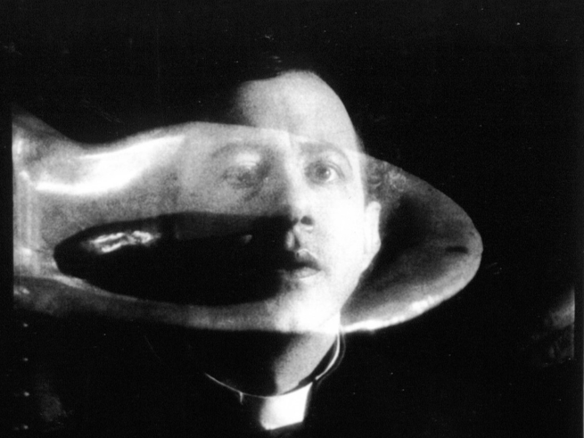 Black and white image of a man in priest garments behind a floating, translucent rounded object.