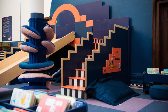 Beautiful shot of the Tiny Totley Studio slide and tactile objects. Colours are dark blue, coral, lilac with black and white stripe stairs up to the slide. Boxes of early years library books to read and borrow.